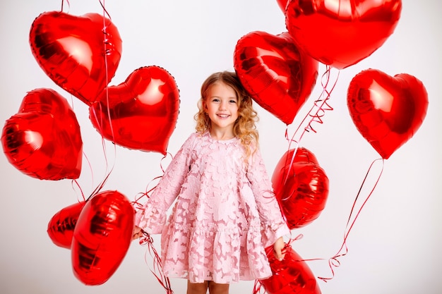 kleines blondes Mädchen in einem rosa Kleid lächelt und hält viele rote herzförmige Luftballons auf einem weißen Wand-Valentinstag-Konzept