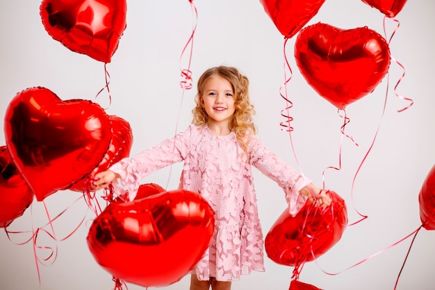 kleines blondes Mädchen in einem rosa Kleid lächelt und hält viele rote herzförmige Luftballons auf einem weißen Wand-Valentinstag-Konzept