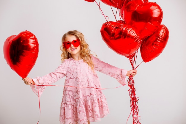 kleines blondes Mädchen in einem rosa Kleid lächelt und hält viele rote herzförmige Luftballons Valentinstagkonzept