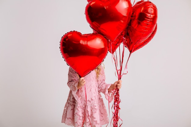 kleines blondes Mädchen in einem rosa Kleid lächelt und hält viele rote herzförmige Luftballons Valentinstagkonzept