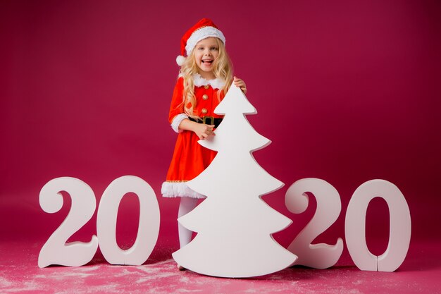 kleines blondes Mädchen in einem Sankt-Anzug hält einen großen weißen Weihnachtsbaum