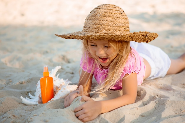 kleines blondes Mädchen in einem Strohhut liegt auf einem Sandstrand neben einer Muschel und Sonnencreme, Platz für Text