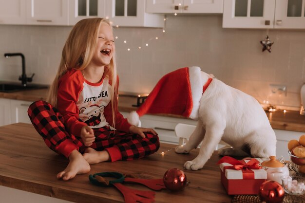 Kleines blondes Mädchen in rotem Weihnachtspyjama in einer Girlandenküche setzt eine Weihnachtsmütze für einen Hund auf. Winterwochenenden und Feiertage im herzlichen Familienkreis. Kind spielt zu Hause mit einem Haustier. Platz für Text