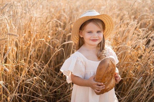 Kleines blondes Mädchen in Strohhut und beigefarbenem Musselinkleid hält Brot im Weizenfeld bei Sonnenuntergang
