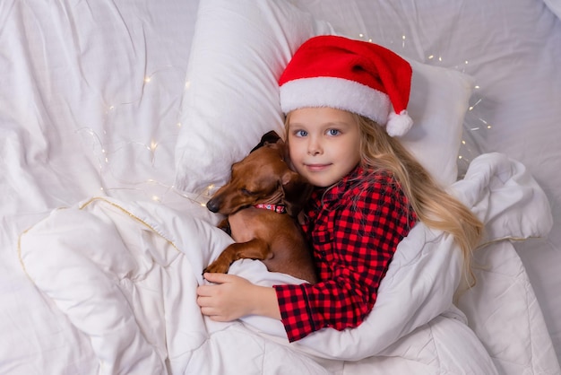 Kleines blondes Mädchen in Weihnachtsmütze und Schlafanzug schläft mit ihrem geliebten Hund Dackel im Bett