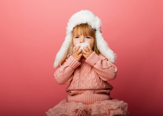 Kleines blondes Mädchen in Winterkleidung und Pelzmütze auf rosa Hintergrund.