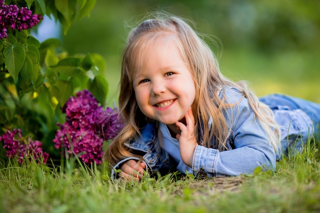Kleines blondes Mädchen liegt in der Nähe der lila Blumen auf dem grünen Gras und lächelt