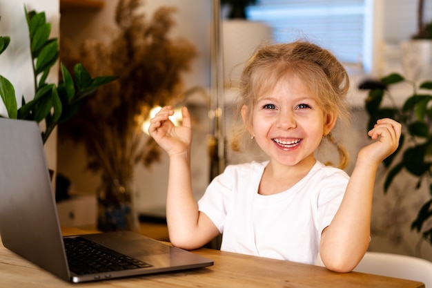 kleines blondes Mädchen mit einem Laptop sitzt an einem Tisch und lächelt