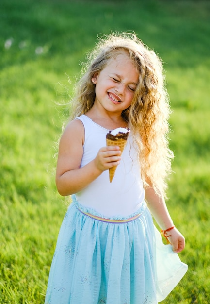 Kleines blondes Mädchen mit langen Haaren hat Spaß beim Eisessen in einem Sommerpark.