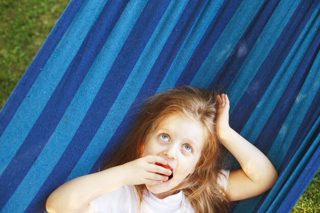 Kleines blondes Mädchen mit langen Haaren isst eine Erdbeere im Garten, die in einer blauen Hängematte liegt