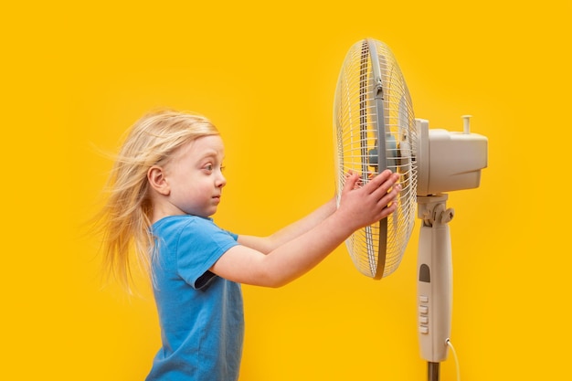 Kleines blondes Mädchen mit vom Wind verwehten Haaren in der Nähe eines Ventilators auf gelbem Hintergrund. Blondes Kind in der Nähe eines Gebläses