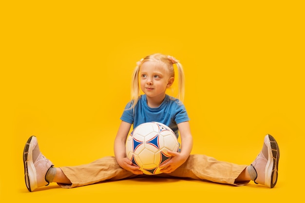 Kleines blondes Mädchen sitzt auf dem Boden und hält Fußball Gelber Hintergrund Frauen-Kinder-Fußball Kinder-Sport