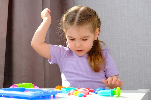 Kleines blondes Mädchen spielt zu Hause mit bunten Plastilinformen auf weißem Tisch