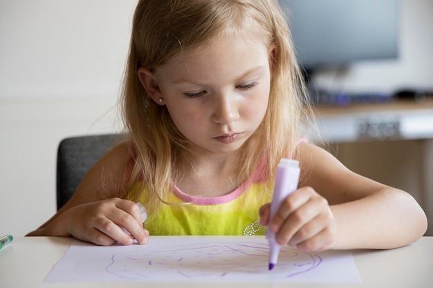 Kleines blondes Mädchen zeichnet, während es am Tisch sitzt