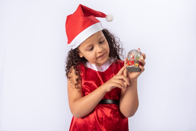 Kleines brasilianisches Mädchen gekleidet mit Weihnachtskostüm, das Schneekugel hält