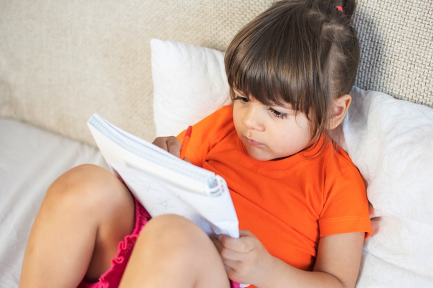 Kleines braunes Haarmädchen mit braunen Augen. 3 Jahre alt. Malen mit Stift im Notizbuch. Zu Hause auf dem Sofa sitzen.