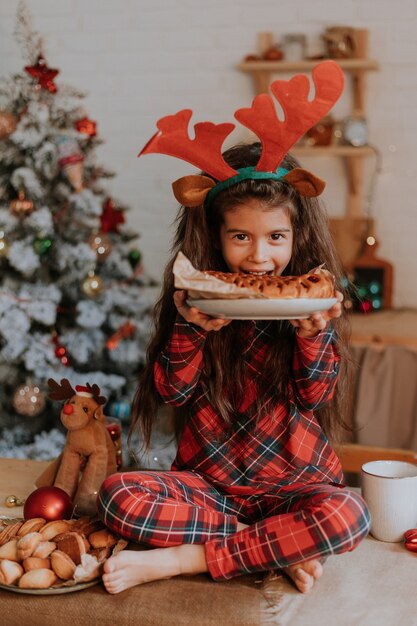 Kleines brünettes Mädchen im karierten roten Pyjama mit Hirschhörnern auf dem Kopf isst einen Weihnachtskuchen