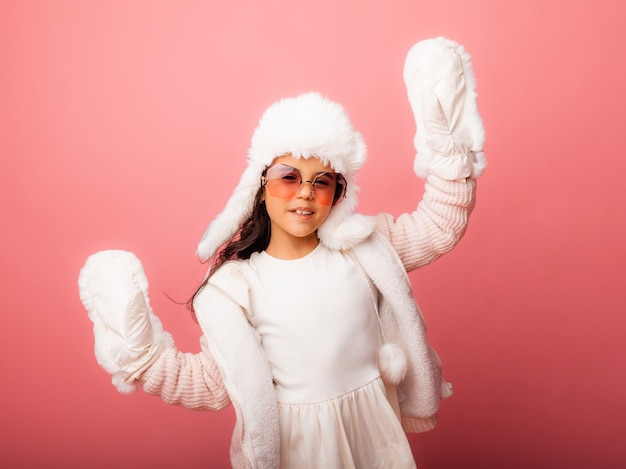 Kleines brünettes Mädchen mit langen Haaren in einer Winterpelzmütze und Fäustlingen auf rosafarbenem Hintergrund.