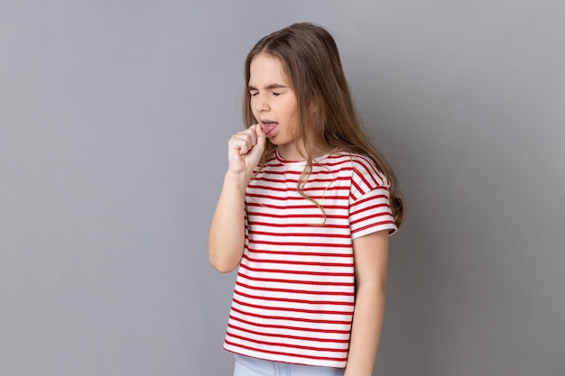 Kleines dunkelhaariges Mädchen mit T-Shirt mit Grippesymptomen, die husten und die Hand in der Nähe des Mundes halten