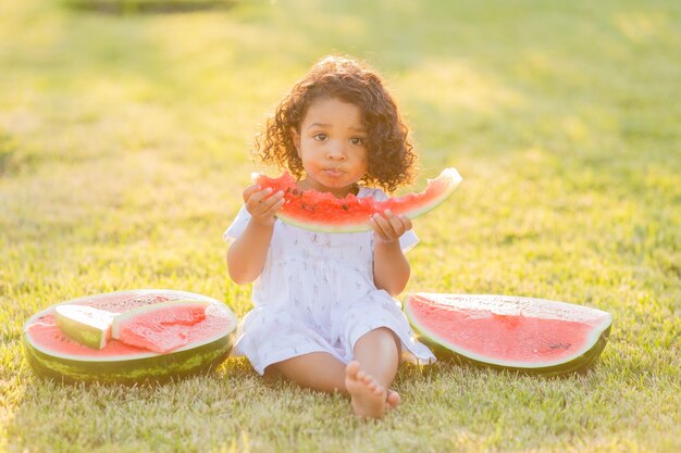 Foto kleines dunkelhäutiges mädchen mit lockigem haar in blassrosa kleid isst eine wassermelone auf dem rasenpicknick im park