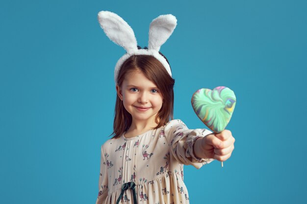 Kleines entzückendes Mädchen, das einen Keks in Form des Herzens hält, der Hasenohren trägt