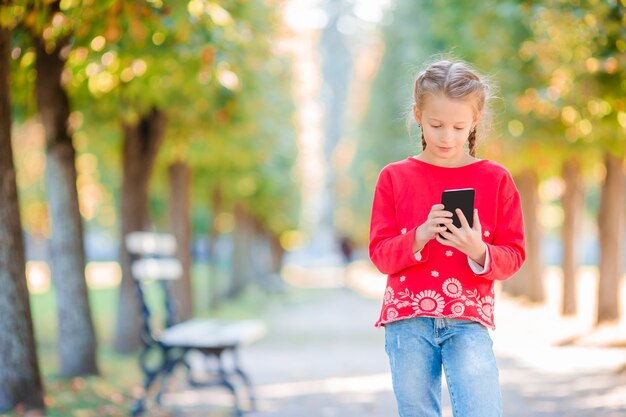 Kleines entzückendes Mädchen mit Smartphone im Fall. Scherzen Sie Spaß am warmen sonnigen Herbsttag draußen haben