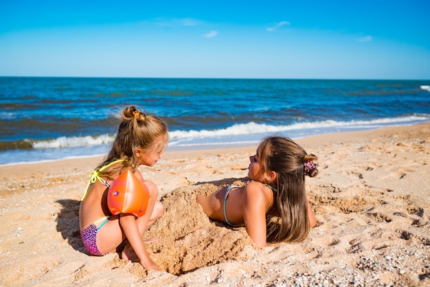 Kleines fröhliches Mädchen begraben ihre ältere Schwester im Sand, während sie an einem sonnigen warmen Sommertag am Strand am Meer spielen