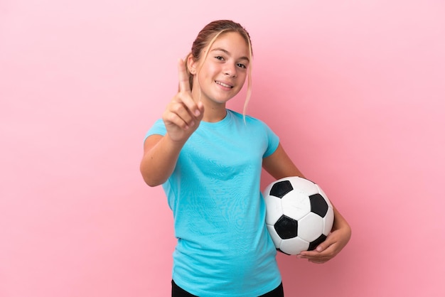 Kleines Fußballspielermädchen isoliert auf rosa Hintergrund, das einen Finger zeigt und hebt