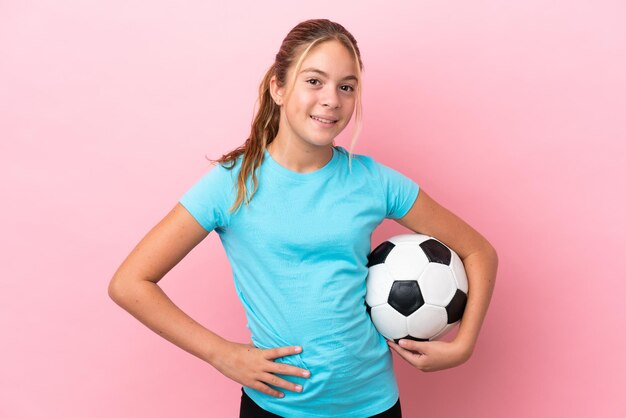 Kleines Fußballspielermädchen isoliert auf rosafarbenem Hintergrund, das mit Armen an der Hüfte posiert und lächelt