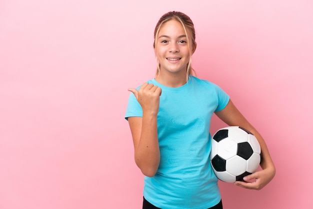 Kleines Fußballspielermädchen isoliert auf rosafarbenem Hintergrund, das zur Seite zeigt, um ein Produkt zu präsentieren