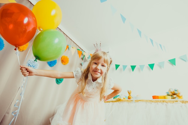 Kleines Geburtstagskind mit Krone auf dem Kopf mit bunten Luftballons. Die Mädchenprinzessin auf der Geburtstagsfeier