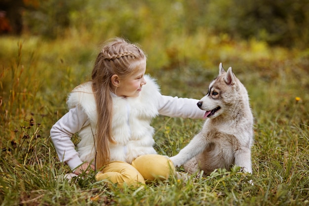 Kleines gelocktes Mädchen sitzt im Herbstwald mit dem Welpenschlittenhund