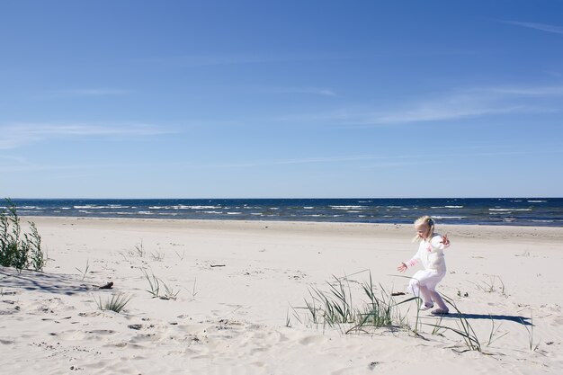 Kleines glückliches blondes Mädchen im weißen Kleid geht am Strand an der Ostseeküste spazieren.