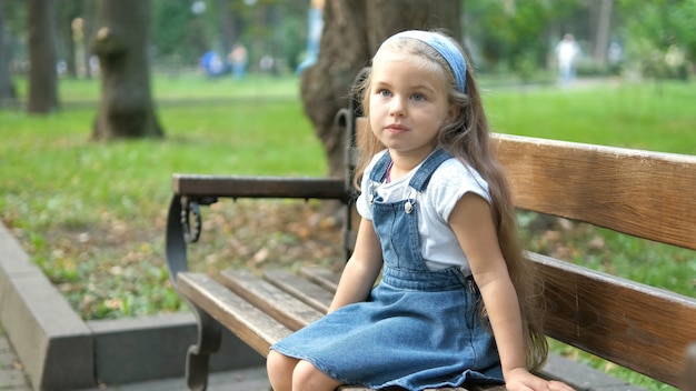 Kleines glückliches Kindermädchen, das auf einer Bank sitzt, die im Sommerpark stillsteht.