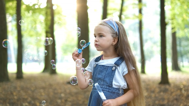 Kleines glückliches Kindermädchen, das draußen im Sommerpark Seifenblasen bläst.
