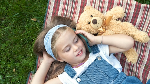 Kleines glückliches Kindermädchen, das im Sommer mit ihrem Teddybären auf dem Handy auf einer Decke auf grünem Rasen liegt.