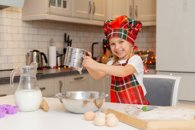 Kleines glückliches lächelndes blondes Mädchen in einem Kochkostüm knetet Teig für Weihnachtsbäckerei in der Küche