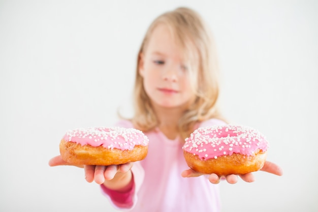 Kleines glückliches Mädchen mit blondem Haar, das Donuts mit rosa Zuckerguss bei Chanukka-Feier spielt und schmeckt