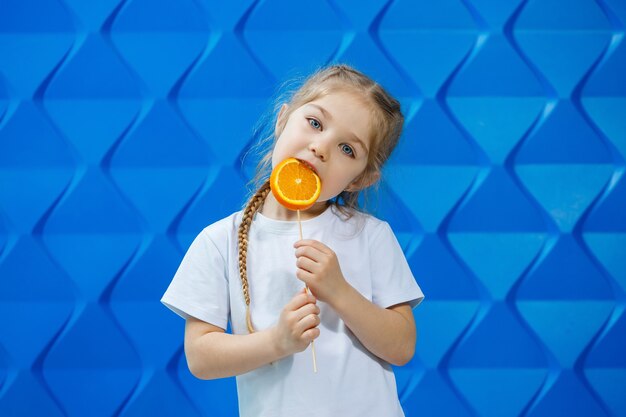 Kleines glückliches mädchen mit einer orange in der hand, schaut in die kamera gekleidet in weißem t-shirt, isoliert auf blauem hintergrund, kopierraum,