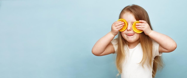 Kleines glückliches Mädchen mit Orangenhälften, die die Augen auf blauem Hintergrund bedecken