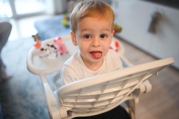 Kleines glückliches süßes Baby Kleinkind Junge Blondine sitzt auf Kinderstuhl und spielt mit Spielzeug Baby Gesichtsbehandlung