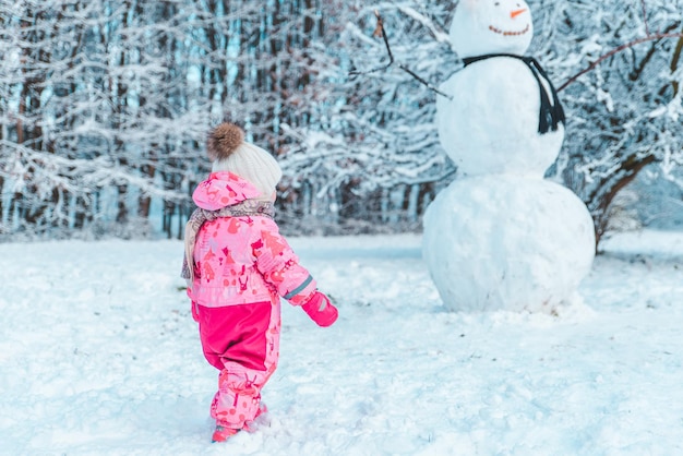 Kleines goldenes Kind in rosa Winterkleidung, das großen Schneemann anschaut