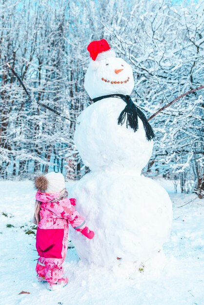 Kleines goldenes Kind in rosa Winterkleidung, das großen Schneemann anschaut