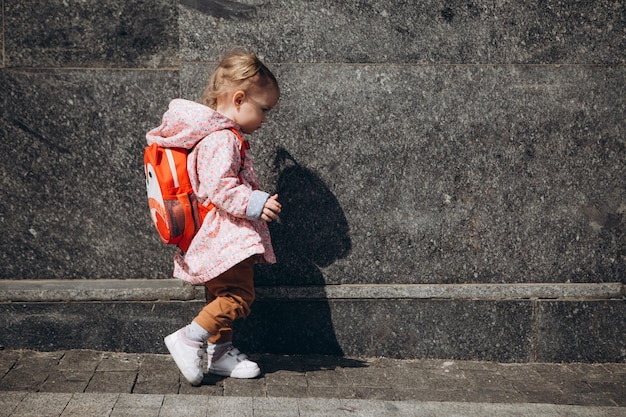 Kleines hübsches Baby, das sich in der Stadt verirrt hat, ein Mädchen in einem rosa Regenmantel mit einem Rucksack