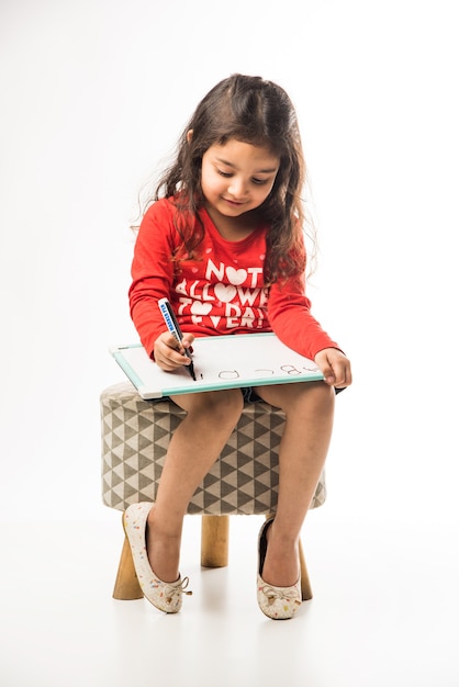 Kleines indisches Mädchen, das mit Filzstift auf Schiefertafel schreibt, während es auf einem Hocker auf weißem Hintergrund sitzt