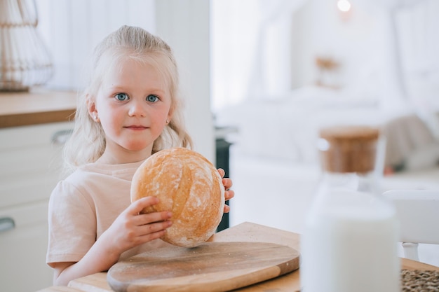 Kleines italienisches Mädchen in legerer Kleidung hält Brot in der Hand und blickt in die Kamera, steht in der Küche. Gesunde Ernährung für Kinder