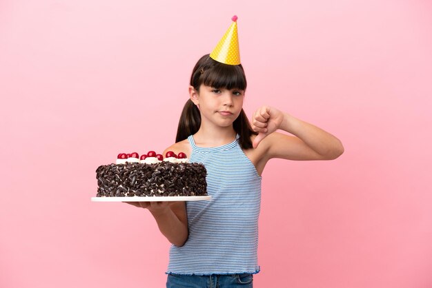 Kleines kaukasisches Kind, das Geburtstagstorte isoliert auf rosafarbenem Hintergrund hält und den Daumen nach unten mit negativem Ausdruck zeigt
