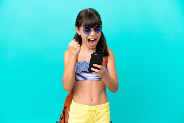Kleines kaukasisches Kind, das isoliert auf blauem Hintergrund mit Telefon in Siegesposition zum Strand geht