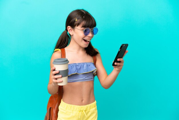 Kleines kaukasisches Kind, das isoliert auf blauem Hintergrund zum Strand geht und Kaffee zum Mitnehmen und ein Handy hält