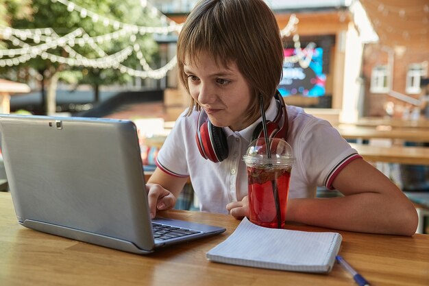 Kleines kaukasisches Mädchen, das am Tisch auf der Veranda des Cafés sitzt und mit Latop studiert